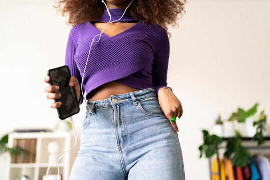 A young woman with curly hair is listening to music through wired headphones, holding her phone. She appears to be enjoying the rhythm, perhaps dancing or simply relaxing to the sound.