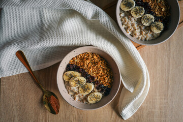 Top view of ceramic bowl filled with oatmeal, sliced bananas, dried cherries, granola and chia seeds, placed on a wooden surface with a spoon and kitchen towel