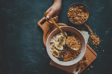 Child’s hand holding a spoon with granola over a bowl of oatmeal with banana slices and dried berries on a dark background. Healthy breakfast concept with dramatic lighting