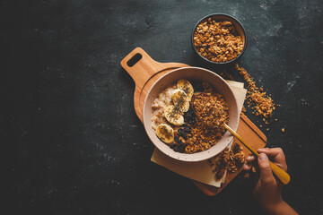 Child’s hand holding a spoon with granola over a bowl of oatmeal with banana slices and dried berries on a dark background. Healthy breakfast concept with dramatic lighting