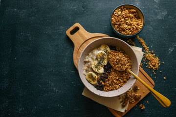Top view of ceramic bowl filled with oatmeal, sliced bananas, dried cherries, granola and chia seeds, placed on a wooden surface with a spoon and kitchen towel