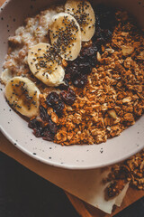 Close-up of healthy oatmeal with banana slices, chia seeds, dried cherries, and granola in ceramic bowl. Nutritious vegetarian breakfast on dark background