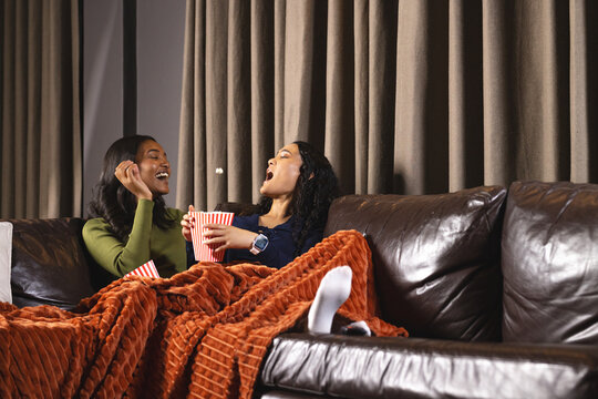 Snacking Diverse female friends sharing orange blanket on sofa at home, tossing popcorn buckets