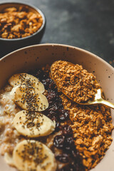 Top view of ceramic bowl filled with oatmeal, sliced bananas, dried cherries, granola and chia seeds, placed on a wooden surface with a spoon and kitchen towel
