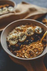 Top view of ceramic bowl filled with oatmeal, sliced bananas, dried cherries, granola and chia seeds, placed on a wooden surface with a spoon and kitchen towel