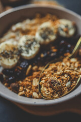 Top view of ceramic bowl filled with oatmeal, sliced bananas, dried cherries, granola and chia seeds, placed on a wooden surface with a spoon and kitchen towel