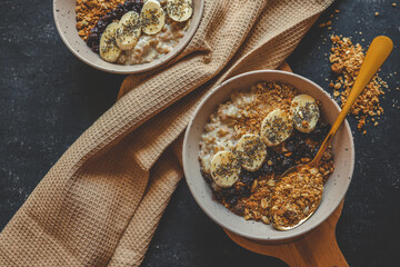 Flat lay of two ceramic bowls with creamy oatmeal, topped with granola, banana slices, dried berries, and chia seeds, placed on a wooden surface with beige waffle towel