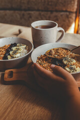 Cozy morning scene with two ceramic bowls of oatmeal topped with banana slices, granola, and chia seeds, next to a mug of tea and a waffle towel