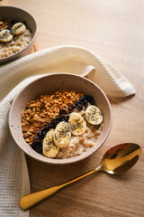 Top view of ceramic bowl filled with oatmeal, sliced bananas, dried cherries, granola and chia seeds, placed on a wooden surface with a spoon and kitchen towel