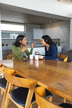 Chatting diverse female friends sitting at open-plan kitchen table, with coffee mugs and smartphone