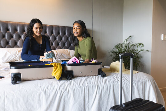 Packing diverse female friends folding colorful clothes on bed at hotel, with suitcase and sun hat