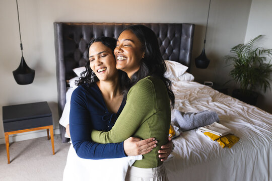 Hugging diverse women standing at foot of bed in bedroom, with rumpled sheets and pendant lamps