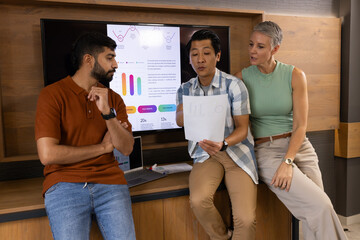Collaborating Diverse colleagues analyzing charts on monitor at counter, with printed data sheet