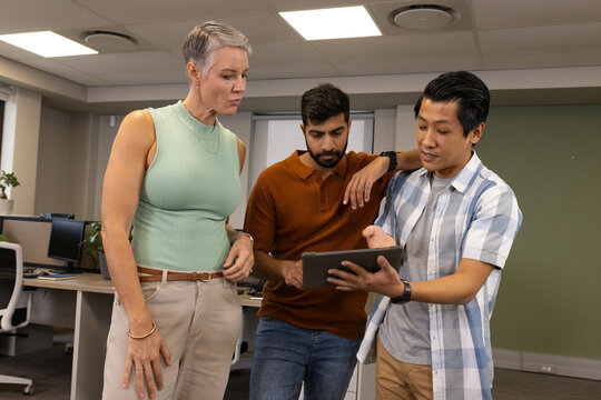 Reviewing Diverse coworkers standing viewing tablet in open-plan office, with computer monitors