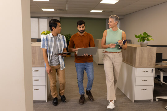 Walking diverse coworkers discussing plans in office corridor, with tablet laptop notebook pen