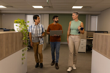 Diverse coworkers walking between cubicles in office aisle, with tablet laptop notebook smartphone