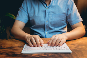 Person in Blue Shirt Typing on White Keyboard in Cozy Workspace with Wooden Table and Green Plant...