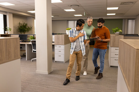 Walking diverse coworkers discussing project in open-plan office corridor, with tablet device