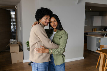 Hugging diverse couple standing in modern open-plan apartment, showcasing moving boxes and plant