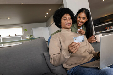 Using laptop Diverse couple sitting on couch at home, holding credit card and mug