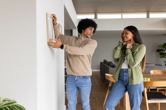 Diverse couple aligning wooden picture frame in open-plan living area, with yellow spirit level