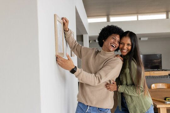 Laughing diverse couple hanging wooden picture frame in modern living room, with tape measure - Powered by Adobe