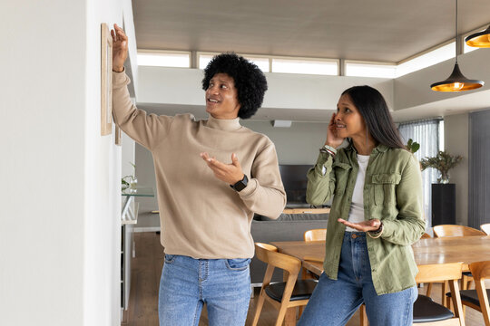 Adjusting wooden picture frame, diverse couple standing in open-plan dining area with pendants