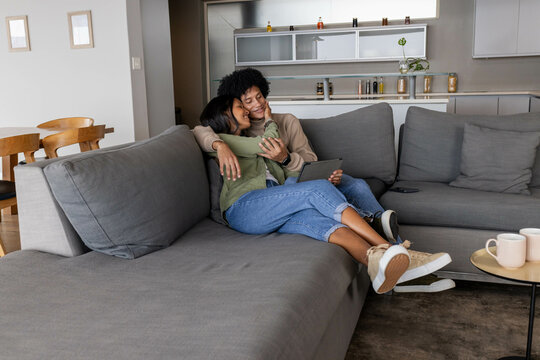 Snuggling diverse couple leaning on gray sectional sofa in living room, with tablet and coffee mugs
