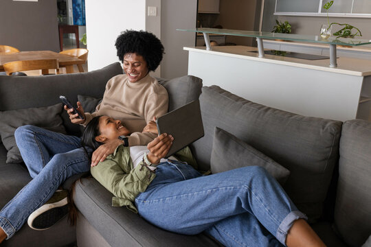 Laughing Diverse couple relaxing on grey sofa in open?plan living room, with smartphone and tablet - Powered by Adobe