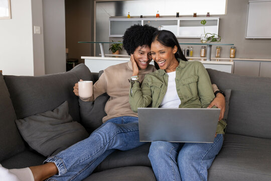 Smiling Diverse couple sitting on grey sofa in open?plan living room, holding coffee mug and laptop - Powered by Adobe
