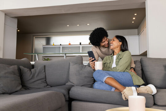 Diverse female partners sharing coffee and smartphone on sofa in modern living room