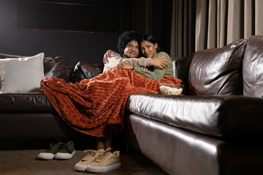 Cuddling diverse couple snuggling under rust-colored blanket on leather sofa at home, with popcorn