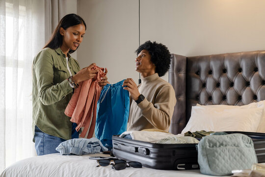 Packing Diverse couple sorting clothes on bed in bedroom, with open black suitcase and pink blouse - Powered by Adobe