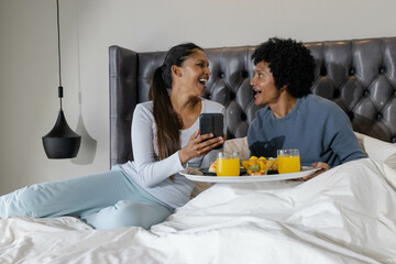 Laughing Diverse couple sitting on bed, holding smartphone and breakfast tray with orange juice