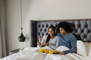 Sharing breakfast tray Diverse couple sitting on bed in bedroom, with orange juice and croissant