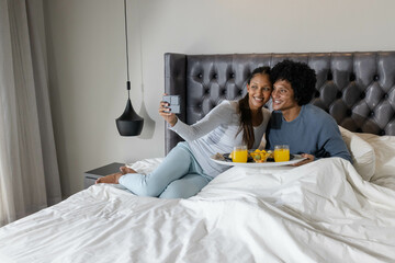 Taking selfie Diverse couple reclining on white bed, with breakfast tray and smartphone