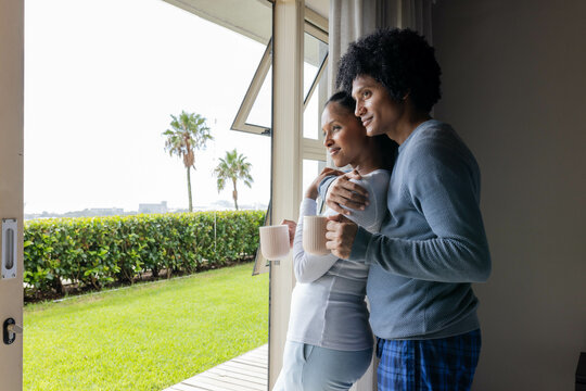 Hugging Diverse couple standing by open sliding glass door at home with ceramic mugs, copy space - Powered by Adobe