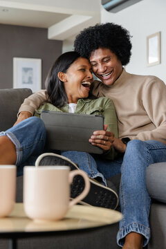 Laughing diverse couple sitting closely on grey sofa in living room, with tablet and ceramic mugs