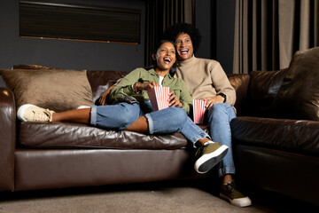 Diverse couple sitting on leather sofa in living room watching movie, with striped popcorn boxes