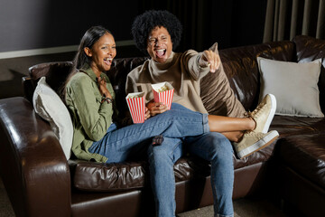 Laughing diverse couple sitting on dark brown leather sofa in home theater, holding popcorn boxes