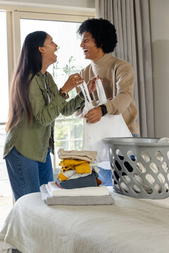 Laughing diverse couple folding laundry in bright bedroom, with grey laundry basket
