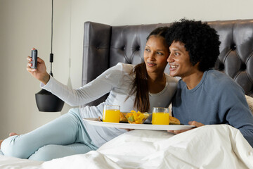 Smiling Diverse couple taking selfie in bedroom, with white breakfast tray and orange juice