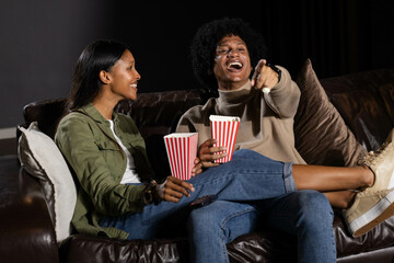 Laughing Diverse couple sitting on brown leather couch in home theater, with striped popcorn boxes