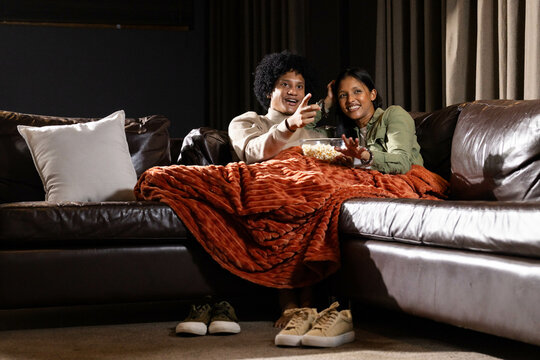 Diverse couple snuggling on sectional couch at home, sharing popcorn and rust-colored throw blanket