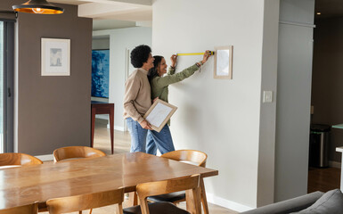 Hanging Diverse couple positioning framed picture on modern dining room wall, using yellow tape