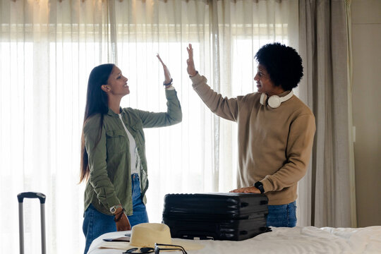 High-fiving diverse couple standing next to black suitcase in hotel room, with straw hat camera