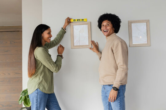 Aligning Diverse couple mounting framed prints on hallway wall, with yellow spirit level and plant - Powered by Adobe