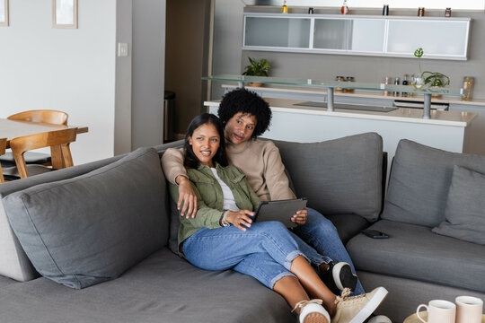 Sitting diverse couple sharing tablet on gray sectional sofa in open plan area, with smartphone - Powered by Adobe