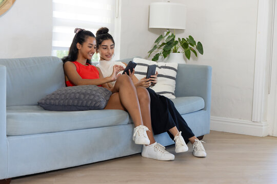 Chatting diverse female friends sitting on light blue sofa in modern living room, with smartphones