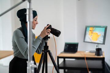 A woman in a headscarf is taking a product photo with a camera on a tripod in a studio setting. She is likely capturing images for e-commerce or marketing purposes, as seen on the monitor display.